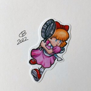 Könnte beinhalten: Eine Cartoon-Illustration eines Mädchens mit blonden Haaren, das ein rosa Kleid und rote Schuhe trägt. Sie hält eine schwarze Bratpfanne in ihrer rechten Hand. Das Bild ist ein Aufkleber mit weißem Hintergrund.