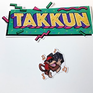 Puede incluir: Dos pegatinas, una con la palabra "TAK KUN" en una fuente retro de los años 80 colorida y la otra con un mono de dibujos animados que lleva gafas de sol y un sombrero rojo, sosteniendo una radiograbadora y rodeado de notas musicales.