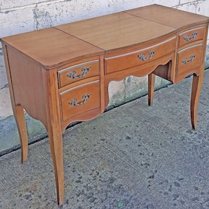 May include: A medium-brown wooden vanity desk featuring a rectangular top with a central flip-top compartment, four side drawers with ornate dark metal handles, and curved legs. The desk has a smooth finish and a scalloped apron design.
