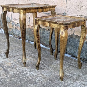 May include: A set of two square side tables with a distressed gold finish. Each table features a flat top with a decorative central motif, scalloped edges, and four slender, curved cabriole legs that taper toward the bottom feet.