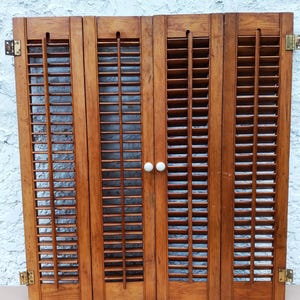 May include: A set of four connected wooden shutters with a medium brown finish and horizontal slats. The shutters feature a vertical central rod on each panel and two small white round knobs on the two center panels, with metal hinges.