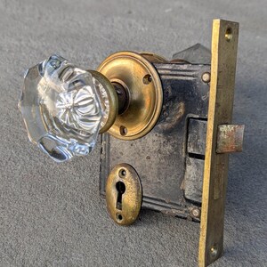 May include: A mortise door lock set featuring a clear, faceted glass knob attached to a circular brass rosette. The mechanism includes a black metal box, a brass faceplate with screw holes, and a separate oval brass keyhole escutcheon plate.