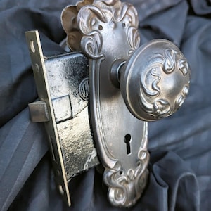 May include: A silver-toned metal door knob set featuring an ornate, embossed scrollwork design on the circular handle and the elongated backplate. The assembly includes a rectangular black metal mortise lock mechanism with a visible latch bolt.