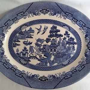 May include: An oval ceramic serving platter featuring a blue and white willow pattern. The central design depicts a landscape with pagodas, a bridge, trees, and two birds in flight. The border is decorated with a geometric lattice pattern.