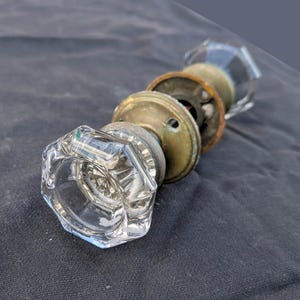 May include: A vintage door knob set featuring two clear, faceted glass knobs with an octagonal shape. The knobs are connected by a central metal spindle and include two circular, weathered brass-toned mounting plates with visible screw holes.