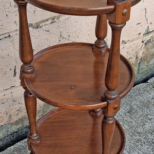 May include: A three-tiered round wooden stand featuring a smooth, polished brown finish. The piece consists of three circular shelves supported by four vertical turned legs with decorative block joints at each level, creating a tiered structure.