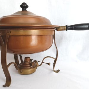 May include: A copper chafing dish with a matching lid, featuring a black wooden handle and a black knob on the lid. The dish is supported by a brass-toned metal stand that includes an integrated burner unit positioned directly beneath the bowl.