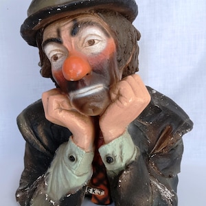 May include: A painted bust of a clown wearing a dark hat, a dark jacket, and a light-colored shirt with a patterned tie. The clown has a bright orange nose, white face paint, and dark eye makeup. The letters EKJ are visible on the tie.