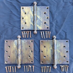 May include: Three rectangular brass door hinges with a weathered patina and ball finials. Each hinge features eight pre-drilled screw holes. A collection of matching metal screws is arranged in small groups below each hinge unit.