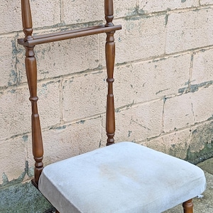 May include: A wooden valet chair featuring a dark brown finish, a built-in hanger at the top, and a horizontal bar for trousers. The seat is upholstered in a light gray fabric, and the chair is supported by four turned wooden legs with dark feet.