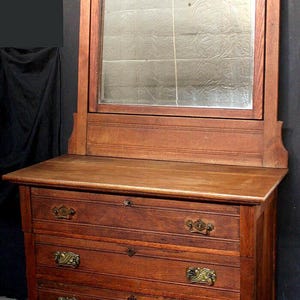 May include: A brown wooden dresser featuring a rectangular mirror attached to the top. The dresser has three drawers with ornate brass handles and keyholes. The mirror frame includes carved floral details at the top corners and center.