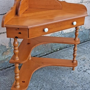 May include: A light brown wooden corner table featuring a triangular top with a raised back edge, a single drawer with two white round knobs, and two lower triangular shelves supported by four turned legs with a smooth, polished finish.
