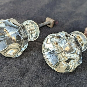 May include: Two clear glass cabinet knobs with a faceted, multi-sided design. Each knob features a reflective, transparent surface and is attached to a short, threaded metal bolt with a square-shaped base for secure installation on furniture.