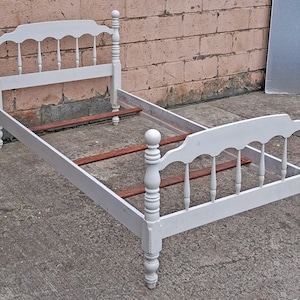 May include: A twin size bed frame painted in a matte light gray finish. The headboard and footboard feature a scalloped top rail, vertical spindle slats, and turned finial posts. Three dark brown wooden support slats span the frame width.