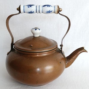 May include: A copper tea kettle with a rounded body and a matching lid. The kettle features a white ceramic handle and a white ceramic lid knob, both decorated with blue floral patterns. The handle is attached to the kettle with metal brackets.
