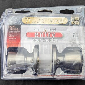May include: A TruGuard stainless steel entry door knobset in clear plastic retail packaging. The set features keyed locking, ANSI grade 3 residential use, universal fit, and includes two keys. The packaging displays a 25-year warranty.