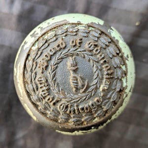 May include: A round metal door knob with a pale green chipped finish. The center features a raised emblem of a hand holding a torch, surrounded by a laurel wreath and the embossed text Board of Education City of Chicago in dark metal.