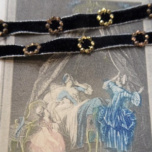 May include: Two black velvet ribbons with gold and brown circular embellishments. The ribbons are on top of a vintage print of a scene with three women in a bedroom. The print has the text "Le Lever" and French text below.