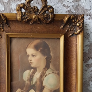 May include: A framed portrait of a young girl with long brown hair wearing a light blue and green dress. The frame is ornate with gold accents and a bow at the top.