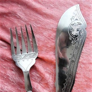 Puede incluir: Un tenedor y un cuchillo de plata con diseños ornamentados y mangos de marfil. Los cubiertos están sobre un fondo de tela rosa.