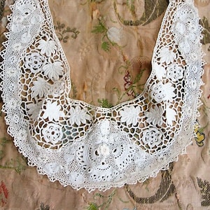 May include: A white crocheted lace collar with a scalloped edge and floral designs. The collar is laid out on a patterned fabric background.