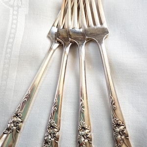 May include: Close-up of several silver forks with decorative floral and leaf patterns on the handles. The forks are fanned out, tines upwards. The background is a white, textured surface. The forks are made of silver.