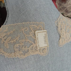 May include: Two pieces of delicate, off-white lace trim with a floral design. The lace is displayed on a light blue fabric background. A small white tag with the text "Carolyn Shop, 362105, New York" is visible.