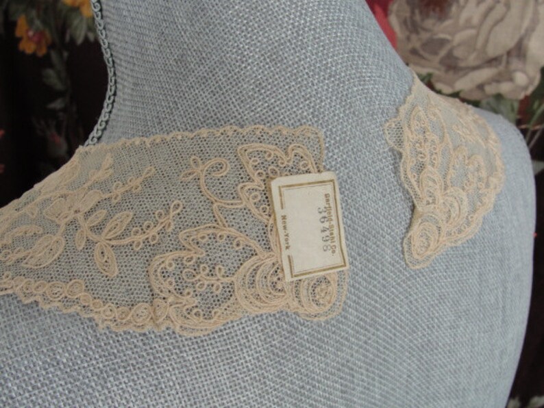 May include: Two pieces of delicate, off-white lace trim with a floral design. The lace is displayed on a light blue fabric background. A small white tag with the text "Carolyn Shop, 362105, New York" is visible.