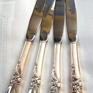 May include: Four silver knives with floral detailing on the handles. The blades are polished and reflect light. Each blade is stamped with the word "COMMUNITY". The knives are displayed on a white surface.
