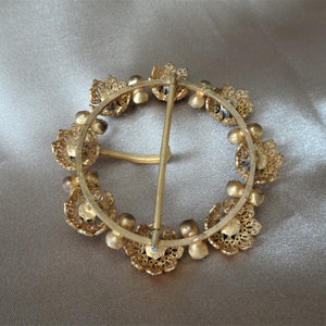 May include: A gold-toned metal belt buckle with a circular design. The buckle is decorated with a ring of small, ornate flowers with blue accents. The buckle has a metal pin and loop closure.