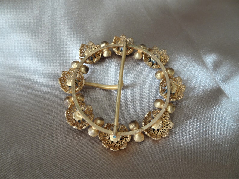 May include: A gold-toned metal belt buckle with a circular design. The buckle is decorated with a ring of small, ornate flowers with blue accents. The buckle has a metal pin and loop closure.
