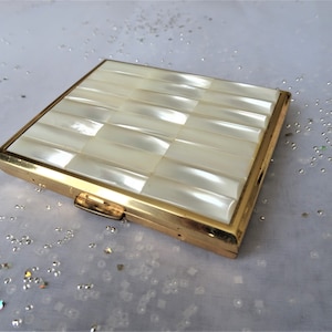 May include: A gold-toned compact with a white mother-of-pearl design. The compact has a hinged lid and a small latch.