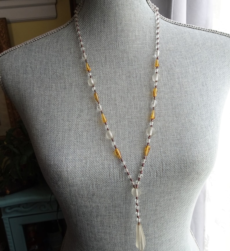 Peut inclure: Un long collier de perles avec des perles de verre transparentes et ambr&eacute;es. Le collier a un pendentif en forme de larme au bas.