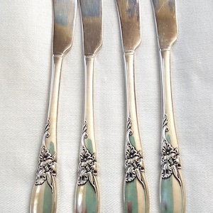 May include: Four silver butter knives with curved blades and floral handle detailing. The knives are displayed vertically against a white backdrop, highlighting their polished surfaces and decorative elements. The blades are approximately 15 cm long.
