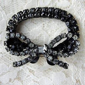 K&ouml;nnte beinhalten: Eine schwarze Strass-Schleifenbrosche mit einem klaren Kristall in der Mitte. Die Brosche besteht aus vielen kleinen Strasssteinen und ist perfekt, um jedem Outfit einen Hauch von Glanz zu verleihen.