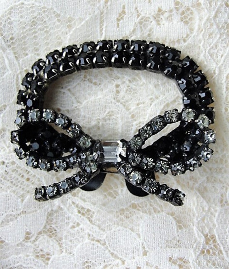 K&ouml;nnte beinhalten: Eine schwarze Strass-Schleifenbrosche mit einem klaren Kristall in der Mitte. Die Brosche besteht aus vielen kleinen Strasssteinen und ist perfekt, um jedem Outfit einen Hauch von Glanz zu verleihen.