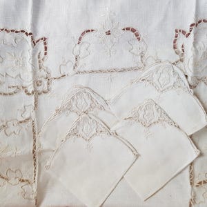 Puede incluir: Un mantel de lino blanco y servilletas a juego. El mantel presenta intrincados calados y diseños florales bordados con un borde marrón claro. Las servilletas tienen un diseño bordado similar. El conjunto es ideal para un comedor formal.