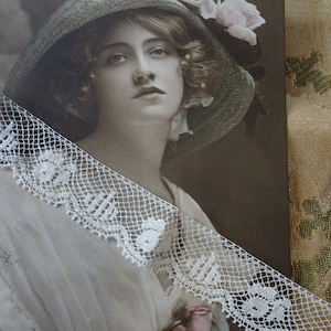 Puede incluir: Una fotografía vintage en sepia de una mujer con un sombrero azul claro con flores rosas. Lleva un cuello de encaje blanco y un vestido blanco. La foto está enmarcada con un borde de encaje blanco.