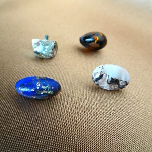 以下が含まれることがあります： 異なる色と模様のガラスボタンが4つあります。ボタンは楕円形で、金属光沢があります。色は青、白、茶色、緑です。