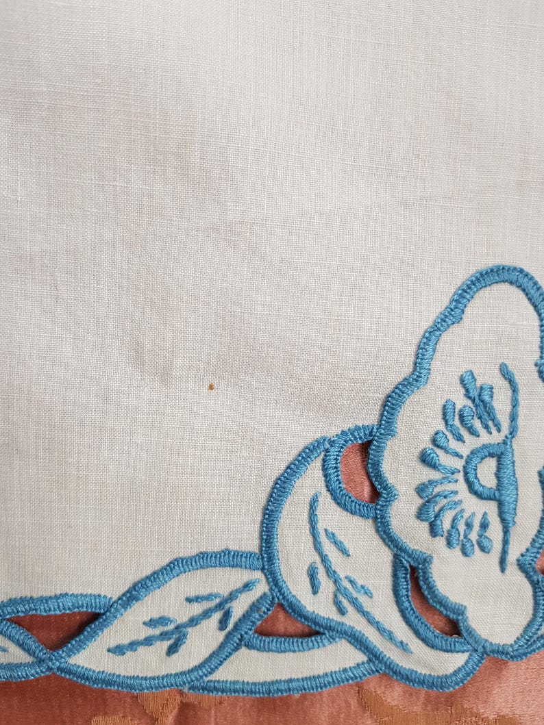 May include: Close-up of a white fabric with a decorative border. The border features a blue embroidered design with floral and leaf motifs. The fabric is set against a peach-colored background.