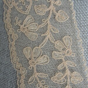 May include: Beige lace trim featuring a delicate floral design. The lace has a scalloped edge and is made from a fine mesh fabric.