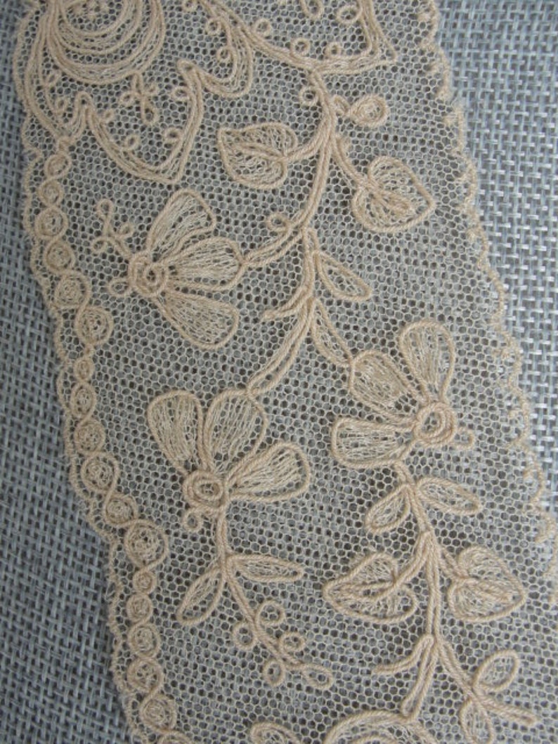 May include: Beige lace trim featuring a delicate floral design. The lace has a scalloped edge and is made from a fine mesh fabric.