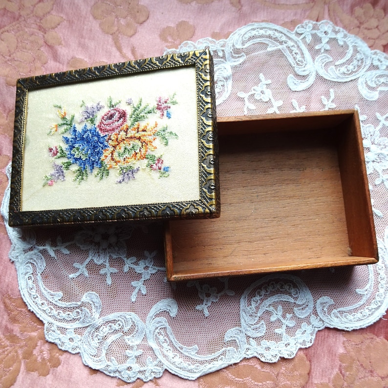 May include: A wooden box with a hinged lid. The lid is decorated with a floral embroidery design featuring a blue, yellow, and pink flower bouquet. The box is open and the inside is empty.