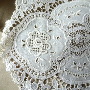 Puede incluir: Primer plano de un tapete blanco bordado con dise&ntilde;os intrincados. El dise&ntilde;o presenta un motivo floral central rodeado de calados delicados y bordes festoneados. Los detalles de encaje crean una est&eacute;tica vintage y artesanal.
