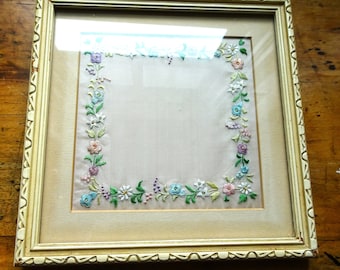 Op de afbeelding: Een wit frame met een delicaat bloemenborduursel in tinten roze, blauw en groen. De borduurwerk is ingelijst met een witte passe-partout en een wit frame.