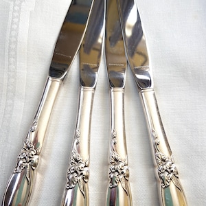 May include: Four silver knives with floral handle detailing. The blades are polished and reflect light. Each blade is marked with the word "COMMUNITY". The knives are displayed on a white surface.