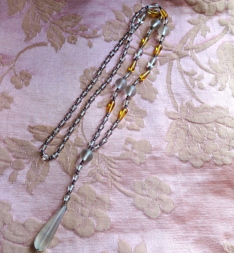 Peut inclure: Un long collier d&eacute;licat compos&eacute; de perles de verre transparentes et ambr&eacute;es. Le collier a un pendentif en forme de larme &agrave; l'extr&eacute;mit&eacute;.