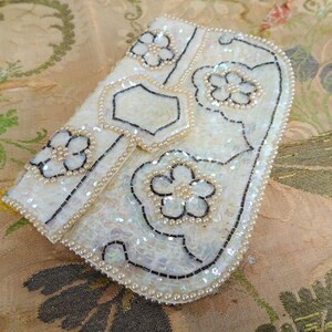 May include: A white beaded clutch purse with a floral design. The purse is made of sequins and pearls and has a black outline.