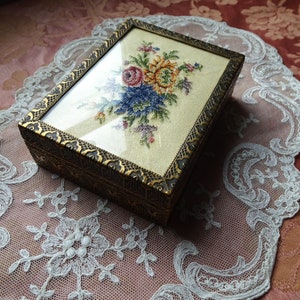 May include: A small, rectangular, wooden box with a glass top. The box is decorated with a gold-colored trim and a floral embroidery design. The embroidery features a bouquet of colorful flowers, including pink, yellow, blue, and green.