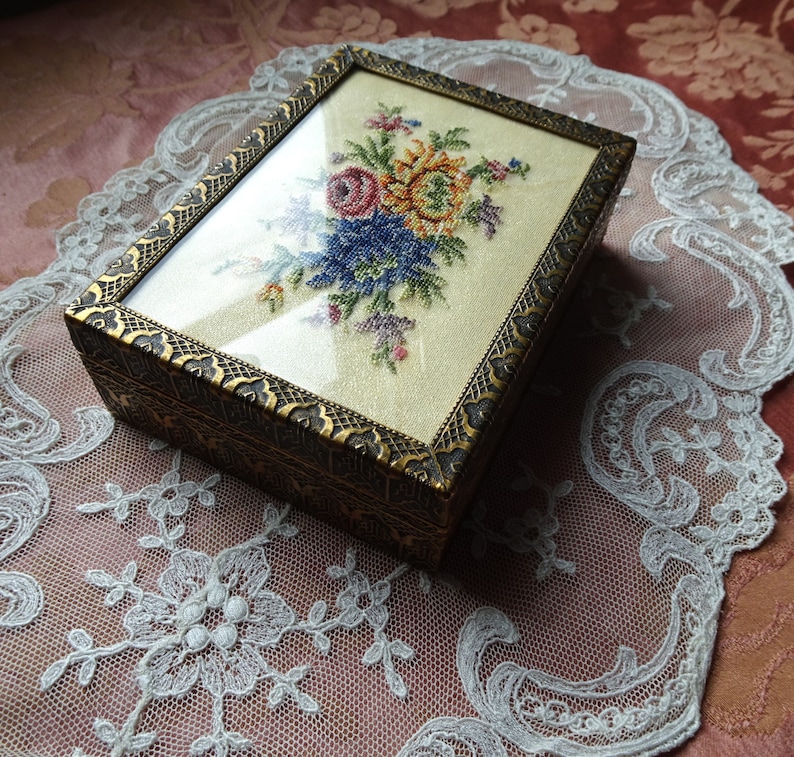 May include: A small, rectangular, wooden box with a glass top. The box is decorated with a gold-colored trim and a floral embroidery design. The embroidery features a bouquet of colorful flowers, including pink, yellow, blue, and green.